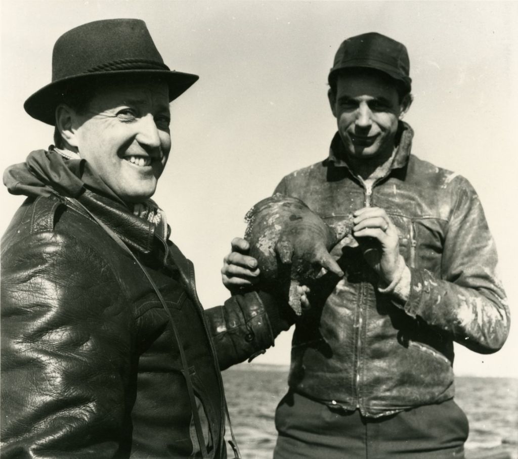 Svartvitt foto av två leende män, en i läderjacka och hatt och den andre i mössa och läderjacka, båda hållandes ett föremål